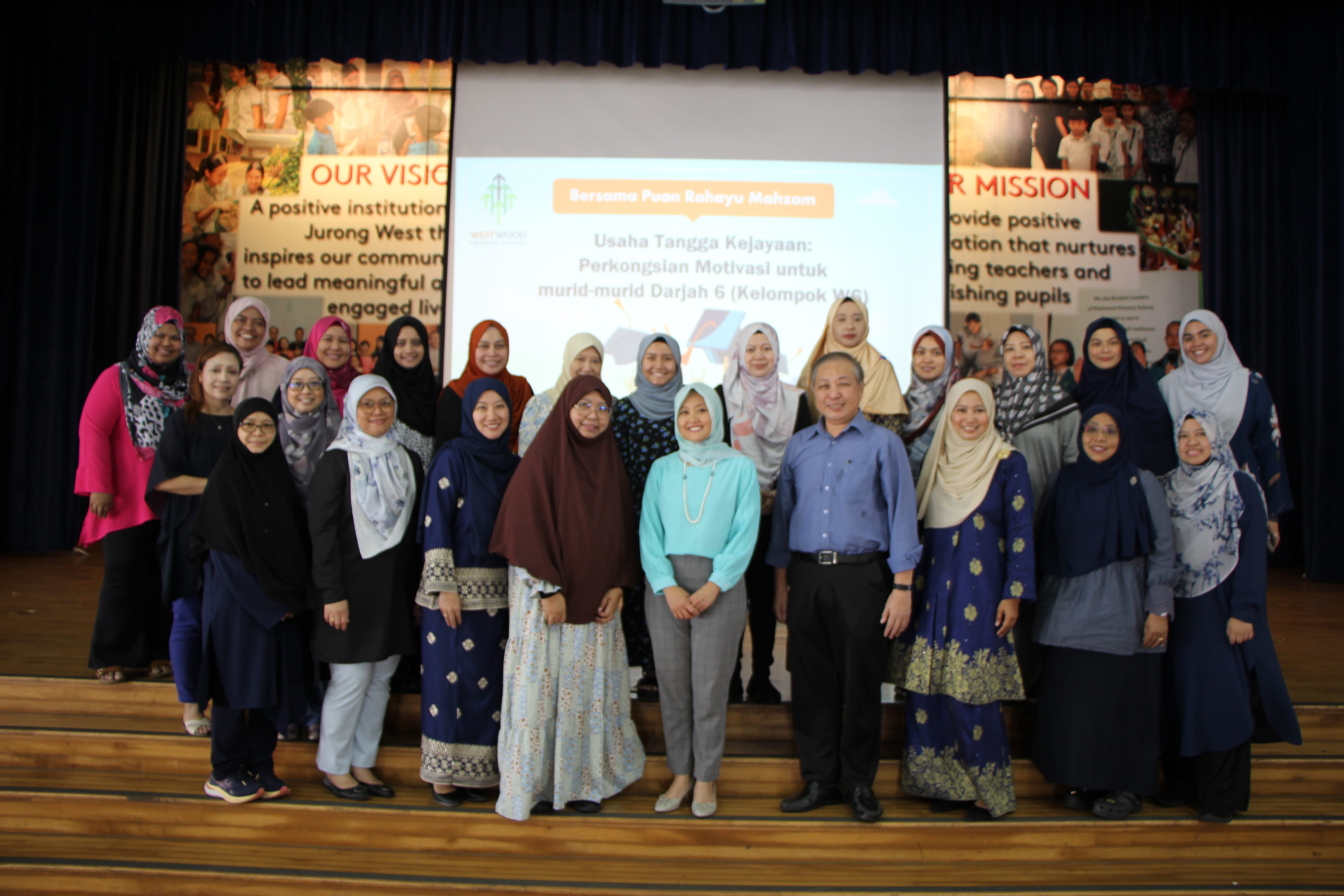 Group photo of staff on a stage in front of a projected presentation screen and school mission posters.